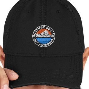 May include: A black distressed baseball cap with a circular patch featuring a mountain range, a sun, and the text "North Goods Co. Get Outdoors".