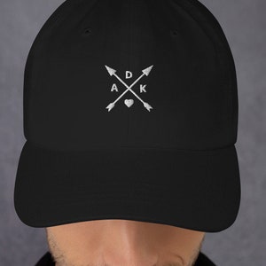 May include: Black baseball cap with a white embroidered design featuring two crossed arrows with a heart and the letters "A D K" in the center.