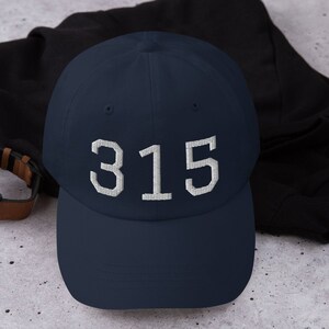 Puede incluir: Una gorra de béisbol azul marino con texto blanco bordado que dice "315".