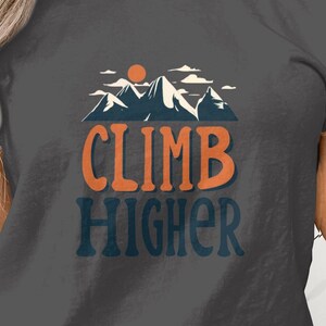 May include: Dark gray t-shirt with a mountain and sun graphic. The shirt features the words "CLIMB HIGHER" in orange and blue lettering. The design includes a mountain range and a setting sun.