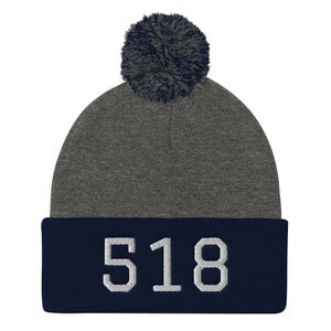 Puede incluir: Gorro de punto gris y azul marino con un pompón en la parte superior. El gorro tiene el número 518 bordado en el puño en blanco.