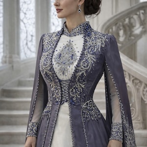 May include: A formal gown with a white bodice and a long, flowing, dark blue overcoat. The gown is embellished with intricate silver and blue beadwork. The sleeves are sheer, and the dress has a high collar. The woman is standing on a marble staircase.