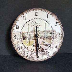 May include: Round clock with a cream-colored face featuring a detailed illustration of Vienna's Schönbrunn Palace. Black hands indicate the time. The clock has black numerals and a decorative outer ring.