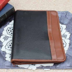May include: A black and brown leather zippered binder. The binder has a textured black surface with a brown accent on the right side. The binder is on a decorative placemat with a white lace doily.