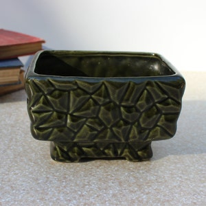 May include: A dark green ceramic planter with a geometric, textured design. The rectangular planter sits on small feet and has a glossy finish. The planter is suitable for small plants and adds a touch of modern style.