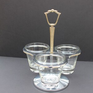 Könnte beinhalten: Ein silberfarbener Gewürzhalter mit drei klaren Gläsern. Der Halter hat einen dekorativen goldfarbenen Griff und einen kreisförmigen Sockel. Die Tassen sind in einem Dreieck angeordnet.