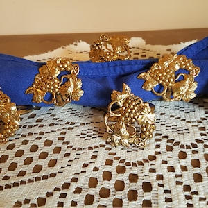 May include: A collection of six gold-colored napkin rings, each featuring a detailed grape and leaf design. The rings are arranged around a folded blue napkin, resting on a white lace tablecloth. The setting suggests a dining or table decor theme.