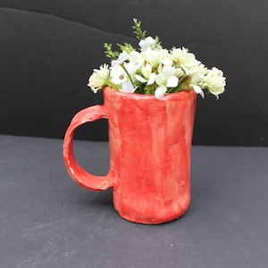 Könnte beinhalten: Eine handgefertigte rote Keramiktasse mit gebogenem Henkel, gefüllt mit künstlichen weißen und grünen Blumen. Die Tasse hat eine strukturierte Oberfläche und steht vor einem dunklen Hintergrund, der ein rustikales, künstlerisches Design zeigt.