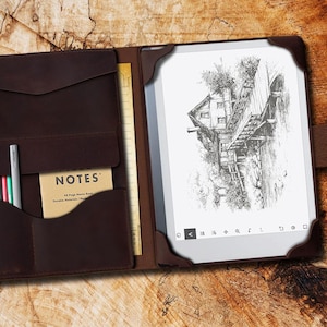 May include: A brown leather notebook cover with a built-in e-reader. The cover has a pocket for pens and a notepad. The e-reader screen shows a black and white drawing of a house with a bridge.