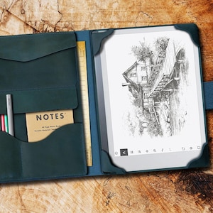 May include: A teal leather notebook cover with a built-in pocket for pens and a notepad. The cover is open to reveal a black and white e-reader with a drawing of a house on a bridge.