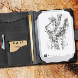May include: A black leather folio case with a built-in notepad and pen holder. The case has a flap that closes with a magnetic snap. The case is open to reveal a black and white e-reader with a drawing of a house on a bridge. The text "NOTES" is visible on the notepad.