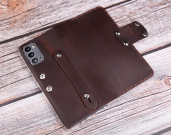 Op de afbeelding: Een donkerbruine leren telefoonhoes met een druksluiting en een handriem. De hoes is ontworpen voor een smartphone en heeft zilverkleurige klinknagels. De hoes is opengeklapt en toont de telefoon.