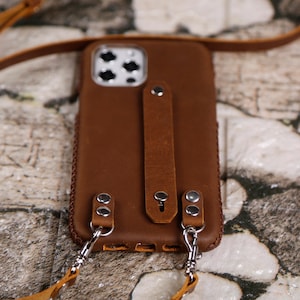 May include: A brown leather phone case with a strap and a silver chain. The case has a built-in hand strap and two metal loops for attaching the chain.