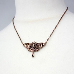 May include: A bronze-toned moth pendant necklace. The pendant showcases a detailed moth design with intricate wing patterns. The fine chain complements the pendant. The necklace is displayed against a white background.
