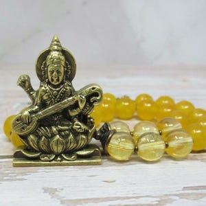 May include: A brass-coloured metal figurine of a seated deity playing a stringed instrument, positioned on a lotus-shaped base. The figurine is accompanied by a strand of translucent yellow beads with a smooth, polished, and spherical finish.