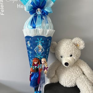 K&ouml;nnte beinhalten: Ein blauer und wei&szlig;er Schult&uuml;te, verziert mit einer blauen Schleife, einem Schneeflockenmuster und zwei Frozen-Puppen. Die Schult&uuml;te steht auf einem Tisch neben einem wei&szlig;en Teddyb&auml;r.