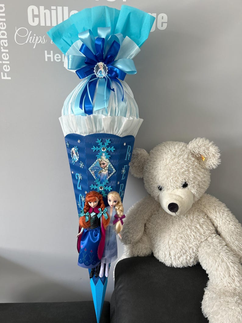 K&ouml;nnte beinhalten: Ein blauer und wei&szlig;er Schult&uuml;te, verziert mit einer blauen Schleife, einem Schneeflockenmuster und zwei Frozen-Puppen. Die Schult&uuml;te steht auf einem Tisch neben einem wei&szlig;en Teddyb&auml;r.
