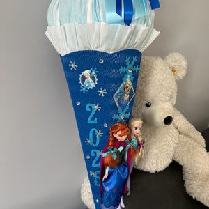 K&ouml;nnte beinhalten: Ein blauer und wei&szlig;er Schult&uuml;te mit einem blauen Band und einer wei&szlig;en Seidenpapier-Oberseite. Die T&uuml;te ist mit Schneeflocken, Strasssteinen und Bildern der Figuren aus dem Film Frozen verziert. Die T&uuml;te tr&auml;gt das Jahr 2023. Zwei Puppen, eine mit roten Haaren und eine mit blonden Haaren, sind an der T&uuml;te befestigt.