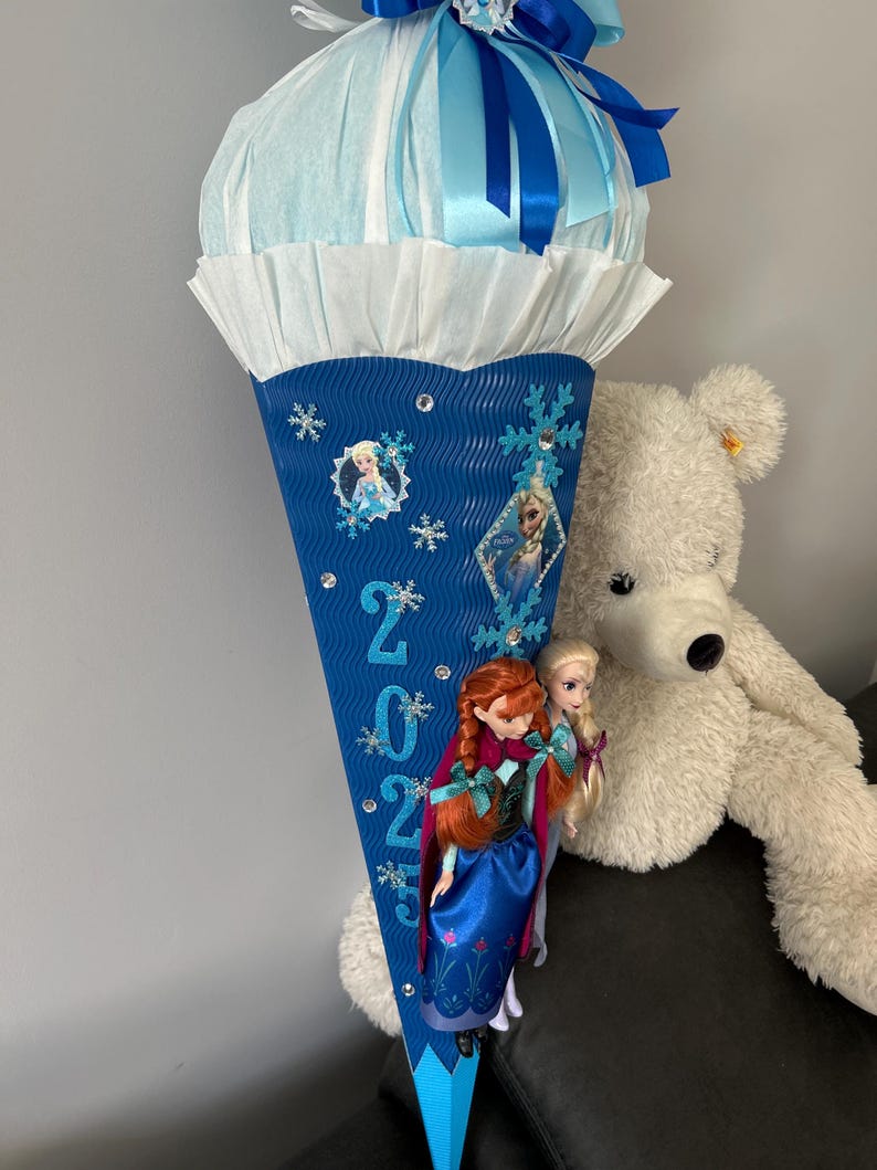 K&ouml;nnte beinhalten: Ein blauer und wei&szlig;er Schult&uuml;te mit einem blauen Band und einer wei&szlig;en Seidenpapier-Oberseite. Die T&uuml;te ist mit Schneeflocken, Strasssteinen und Bildern der Figuren aus dem Film Frozen verziert. Die T&uuml;te tr&auml;gt das Jahr 2023. Zwei Puppen, eine mit roten Haaren und eine mit blonden Haaren, sind an der T&uuml;te befestigt.