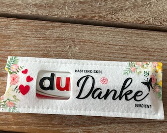 Peut inclure: Un emballage rectangulaire blanc avec le mot "du" en rouge et blanc, et le mot "Danke" en écriture noire. L'emballage est décoré de motifs floraux et de cœurs. Le texte "HAST EIN DICKES" et "VERDIENT" est également visible.