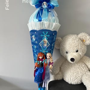 K&ouml;nnte beinhalten: Ein blauer und wei&szlig;er Schult&uuml;te, verziert mit einer blauen Schleife, Schneeflocken und einem Frozen-Motiv. An der T&uuml;te sind zwei Puppen befestigt, eine mit roten Haaren und eine mit blonden Haaren. Die T&uuml;te steht auf einem Tisch neben einem wei&szlig;en Teddyb&auml;r.