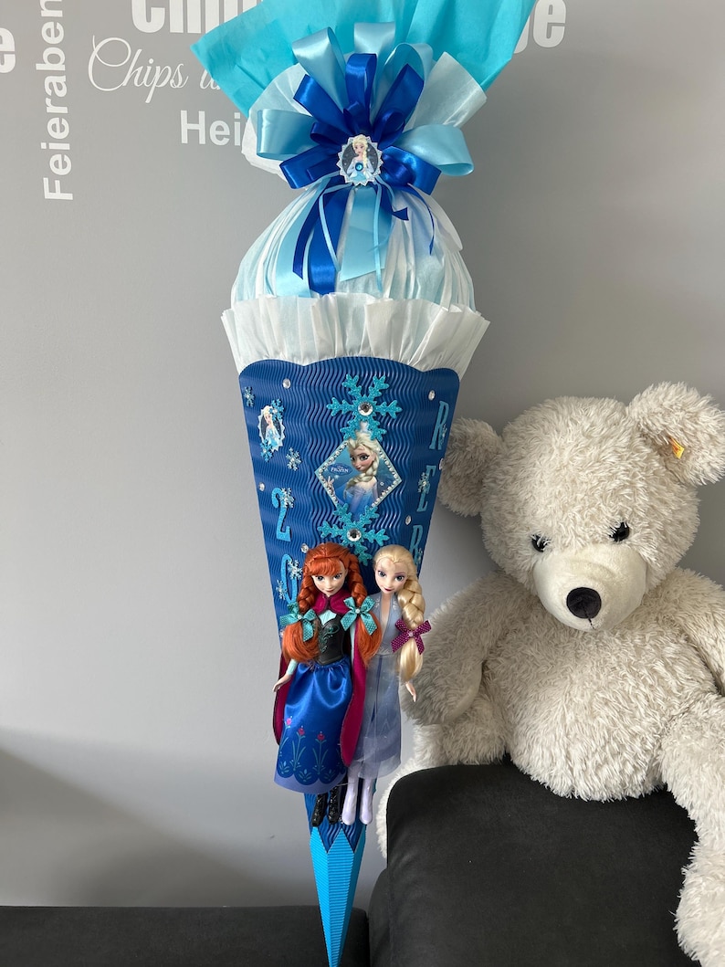 K&ouml;nnte beinhalten: Ein blauer und wei&szlig;er Schult&uuml;te, verziert mit einer blauen Schleife, Schneeflocken und einem Frozen-Motiv. An der T&uuml;te sind zwei Puppen befestigt, eine mit roten Haaren und eine mit blonden Haaren. Die T&uuml;te steht auf einem Tisch neben einem wei&szlig;en Teddyb&auml;r.