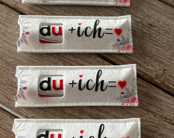 May include: Four white candy bar wrappers with the text "du + ich =" and a red heart. The wrappers have a floral design on the edges and a cutout to show the candy bar inside.