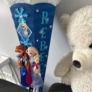 K&ouml;nnte beinhalten: Eine blaue und wei&szlig;e, kegelf&ouml;rmige Schult&uuml;te mit Frozen-Motiv. Die T&uuml;te ist mit Schneeflocken, Glitzer und Strasssteinen verziert. Der Name "Rebecca" ist in blauen Buchstaben auf der T&uuml;te geschrieben. Zwei Frozen-Puppen sind an der T&uuml;te befestigt.