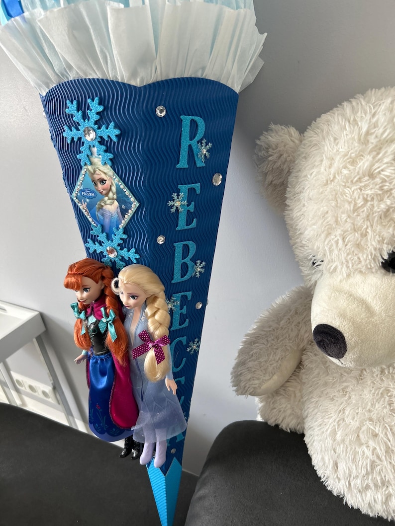 K&ouml;nnte beinhalten: Eine blaue und wei&szlig;e, kegelf&ouml;rmige Schult&uuml;te mit Frozen-Motiv. Die T&uuml;te ist mit Schneeflocken, Glitzer und Strasssteinen verziert. Der Name "Rebecca" ist in blauen Buchstaben auf der T&uuml;te geschrieben. Zwei Frozen-Puppen sind an der T&uuml;te befestigt.