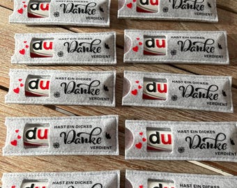 May include: A collection of gray felt candy holders with red heart accents. Each holder contains a candy bar with the word "du" and the words "Hast ein dickes Danke verdient" in black script.