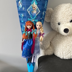 K&ouml;nnte beinhalten: Ein blauer und wei&szlig;er, kegelf&ouml;rmiger Papier-Party-Favorit mit einem Bild von Elsa aus Frozen. Der Kegel ist mit Schneeflocken und Strasssteinen verziert. Zwei Puppen, Anna und Elsa aus Frozen, h&auml;ngen am unteren Ende des Kegels.