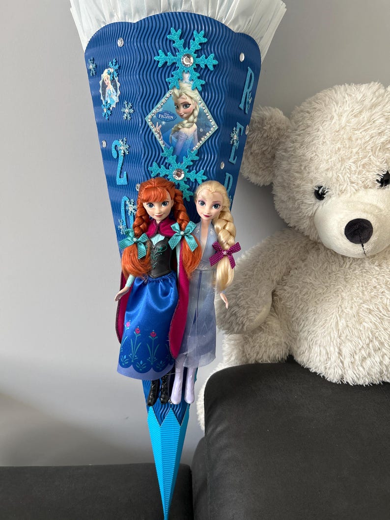 K&ouml;nnte beinhalten: Ein blauer und wei&szlig;er, kegelf&ouml;rmiger Papier-Party-Favorit mit einem Bild von Elsa aus Frozen. Der Kegel ist mit Schneeflocken und Strasssteinen verziert. Zwei Puppen, Anna und Elsa aus Frozen, h&auml;ngen am unteren Ende des Kegels.