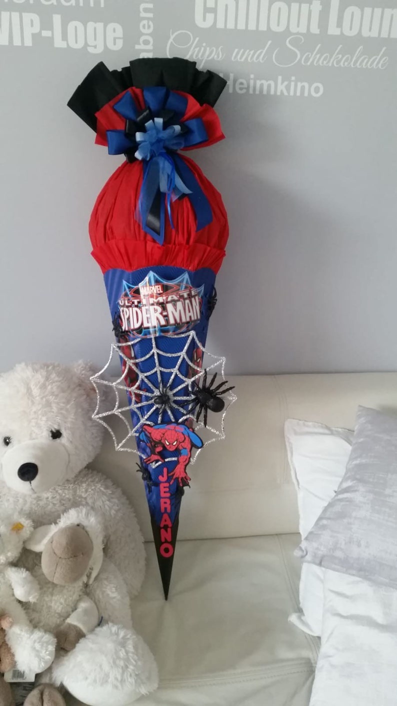 May include: A red and blue school cone with a Spiderman theme. The cone is decorated with a black bow, a silver spider web, and a black spider. The cone is labeled "Ultimate Spiderman" and "Jerano".