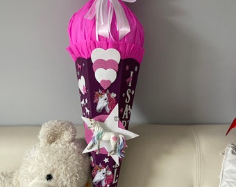 May include: A pink and purple school cone with a unicorn theme. The cone is decorated with a white bow, hearts, stars, and a unicorn figurine. The cone is sitting on a white couch.