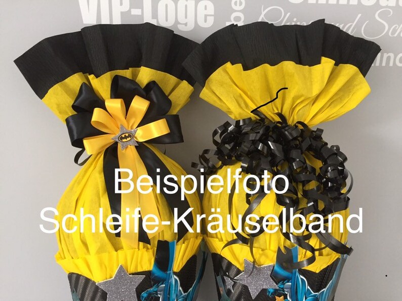 May include: Two yellow and black party cones with a Batman theme. One cone has a black and yellow bow with a Batman charm. The other cone has black curly ribbon. The cones are decorated with silver stars and a Batman graphic. The text "Beispielfoto Schleife-Kr&auml;uselband" is visible on the cones.