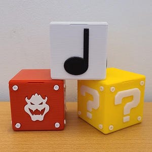 Peut inclure: Trois blocs imprimés en 3D empilés les uns sur les autres. Le bloc du haut est blanc avec une note de musique noire. Le bloc du milieu est rouge avec une tête de Bowser blanche. Le bloc du bas est jaune avec un point d'interrogation blanc.