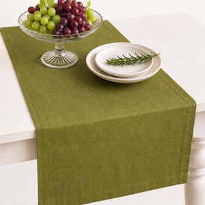May include: A solid olive green rectangular table runner with a stitched hem border. A clear glass pedestal bowl filled with green and red grapes sits on the runner, alongside two stacked white plates topped with a single sprig of green rosemary.