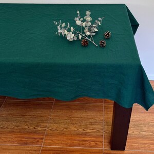 Pode incluir: Uma toalha de mesa de linho verde escuro com um ramo de eucalipto e pinhas no topo. A toalha de mesa está drapeada sobre uma mesa de madeira.