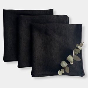 Peut inclure: Trois serviettes en lin noir pliées et empilées sur un fond blanc. Une brindille d'eucalyptus est placée sur le dessus des serviettes.