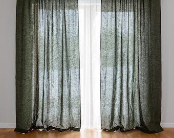 May include: A pair of dark green linen curtains hang in a window, with a white sheer curtain behind them. The curtains are closed, and the light from outside is shining through the sheer curtain.