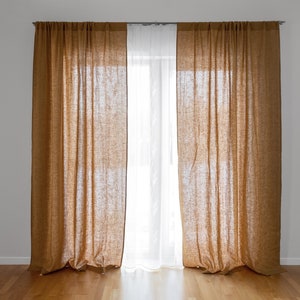 以下が含まれることがあります： 白いシアーカーテンの後ろに、茶色のリネンカーテンが2枚、窓にかけられています。