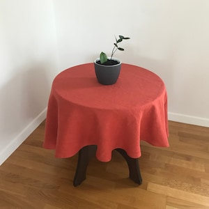 May include: A round, red linen tablecloth draped over a small, dark wooden table. A small potted plant with green leaves sits on the table.