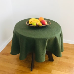 Könnte beinhalten: Eine grüne Tischdecke aus Leinen bedeckt einen runden Tisch mit einem Holzfuß. Ein geflochtener Korb gefüllt mit Obst steht in der Mitte des Tisches.