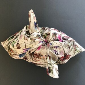 Könnte beinhalten: Eine weiße Stofftasche mit einem Blumendruck in verschiedenen Schattierungen von Rosa, Lila und Grün. Die Tasche ist zu einer dreieckigen Form mit einem Griff gefaltet.