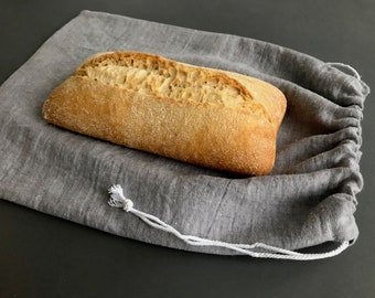 Puede incluir: Una hogaza de pan crujiente descansa dentro de una bolsa de pan gris de lino con cordón. El pan tiene una corteza dorada y una superficie texturizada. La bolsa está hecha de una tela suave y tejida y tiene un cierre de cordón blanco. El fondo es oscuro.