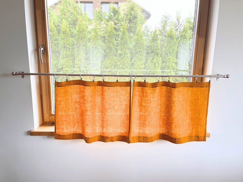 K&ouml;nnte beinhalten: Zwei orangefarbene Vorh&auml;nge aus Leinen, die an einer silbernen Gardinenstange vor einem Fenster h&auml;ngen.