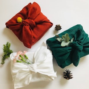 May include: Three fabric-wrapped gifts with natural decorations. A white gift is wrapped with a sprig of pink flowers, a red gift is wrapped with a yellow flower, and a green gift is wrapped with a sprig of eucalyptus.