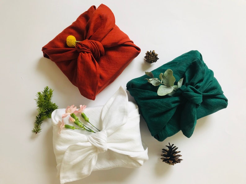 May include: Three fabric-wrapped gifts with natural decorations. A white gift is wrapped with a sprig of pink flowers, a red gift is wrapped with a yellow flower, and a green gift is wrapped with a sprig of eucalyptus.