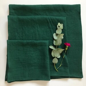 May include: Three green linen napkins of different sizes, folded and stacked on a white surface. A sprig of eucalyptus and a pink carnation are placed on top of the napkins.