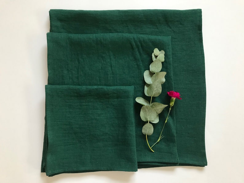 May include: Three green linen napkins of different sizes, folded and stacked on a white surface. A sprig of eucalyptus and a pink carnation are placed on top of the napkins.
